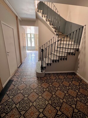 Ein Flur mit gemusterten Bodenfliesen, eine Treppe mit Metallgeländer und eine Tür, die zu einem Außenbereich führt.