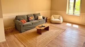 A living room with a sofa, a coffee table, a chair, and a large window with a view of greenery. - 90-square-meter air-conditioned vacation rental / Pool & Garden / Near the beaches (Fleury)
