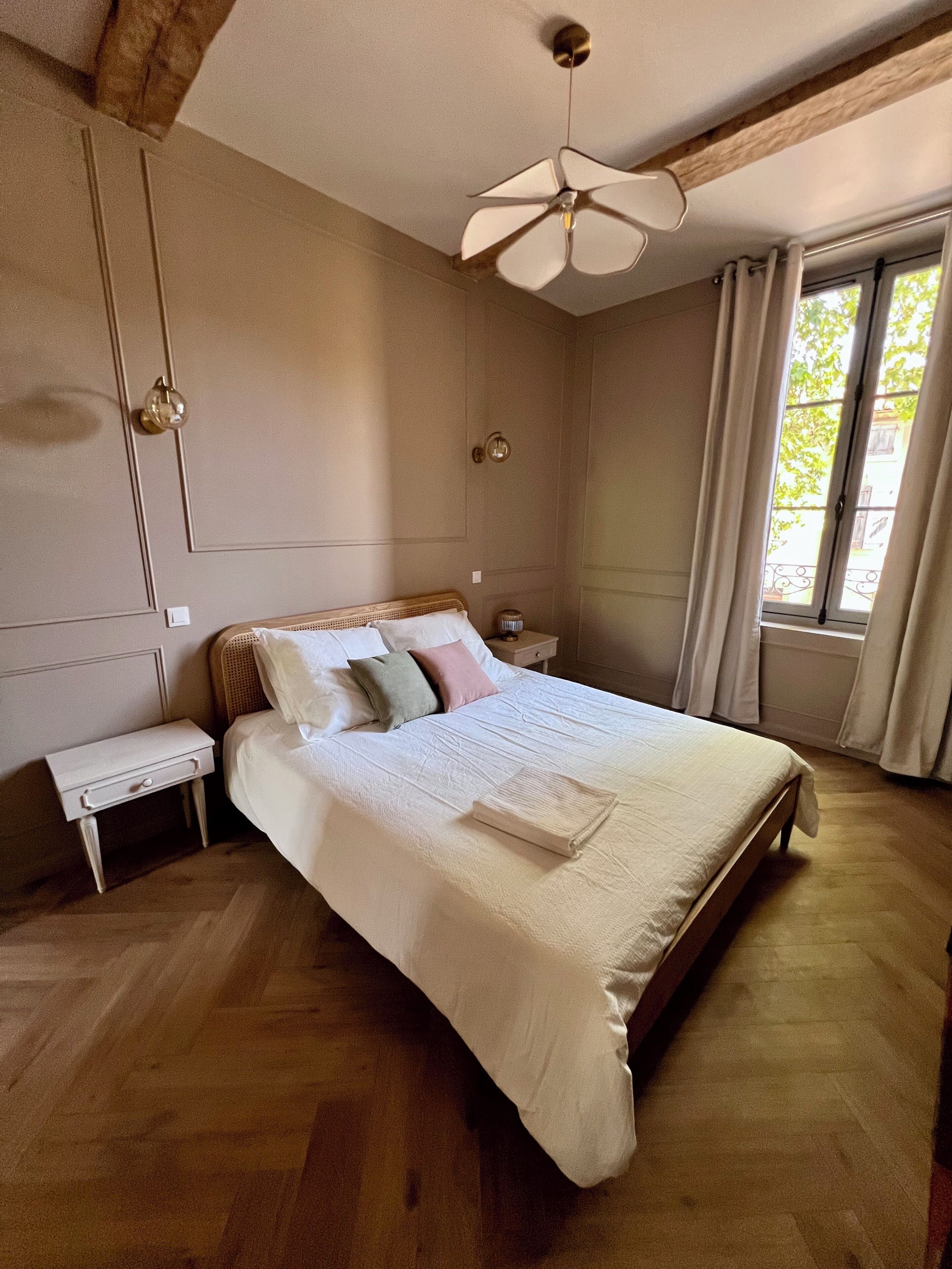 Un dormitori amb un llit gran, una nitzeta, un ventilador de sostre i una finestra amb cortines.