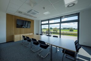 Un salón de conferencias con una larga mesa, sillas negras, un televisor en la pared y amplias ventanas con vistas al exterior.