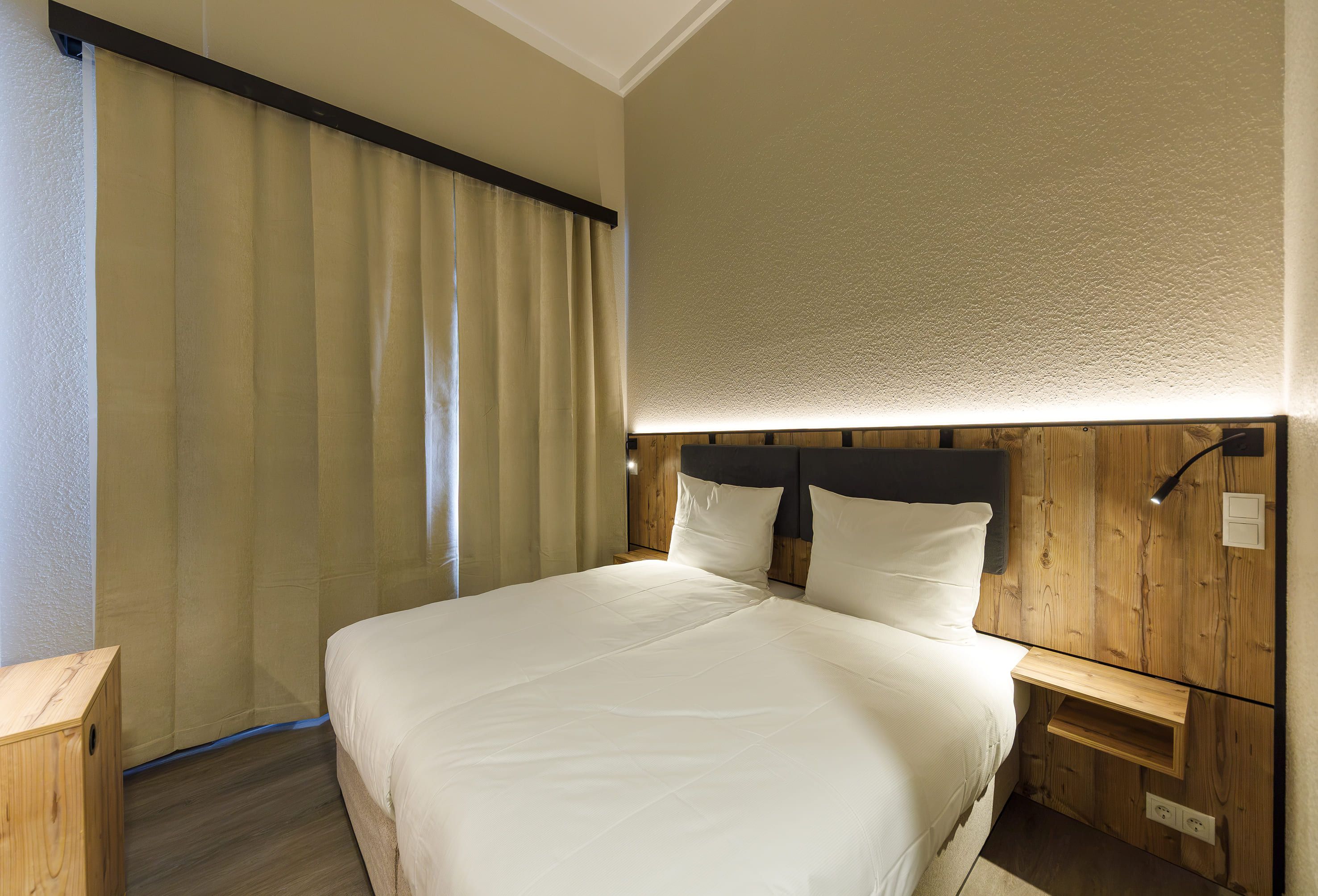 Quarto de hotel com cama, cabeceira de madeira, mesas de cabeceira e cortina.
