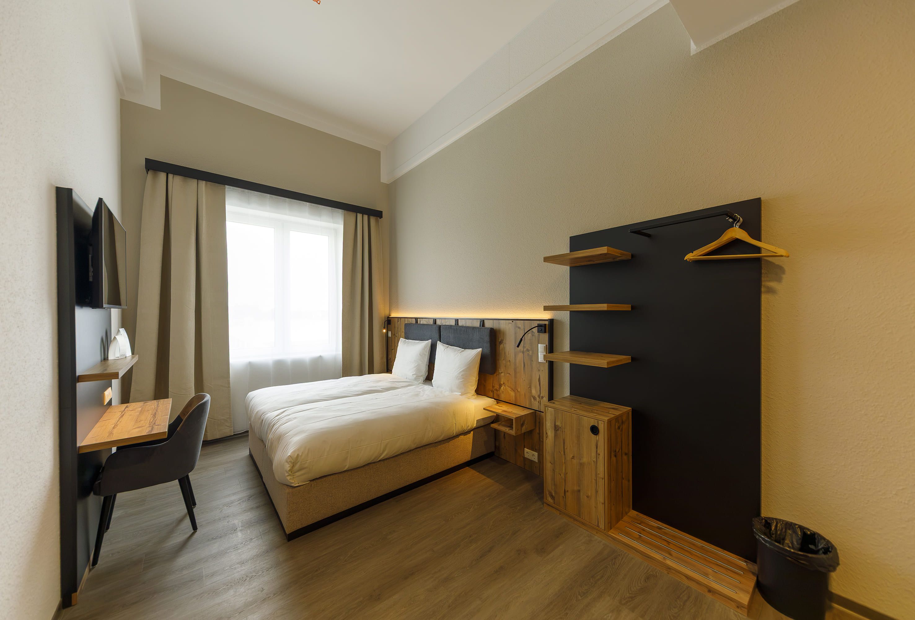 Une chambre d’hôtel comprenant un lit, un bureau, une chaise, une armoire avec des étagères et une fenêtre avec des rideaux.