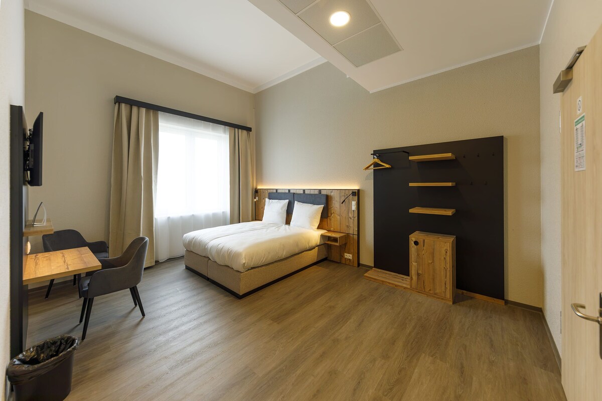A hotel room with a bed, bedside table, lamp, chair, desk, and a TV.