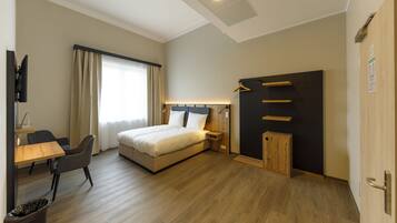 Una habitación de hotel con cama, mesita de noche, lámpara, silla, escritorio y televisión.