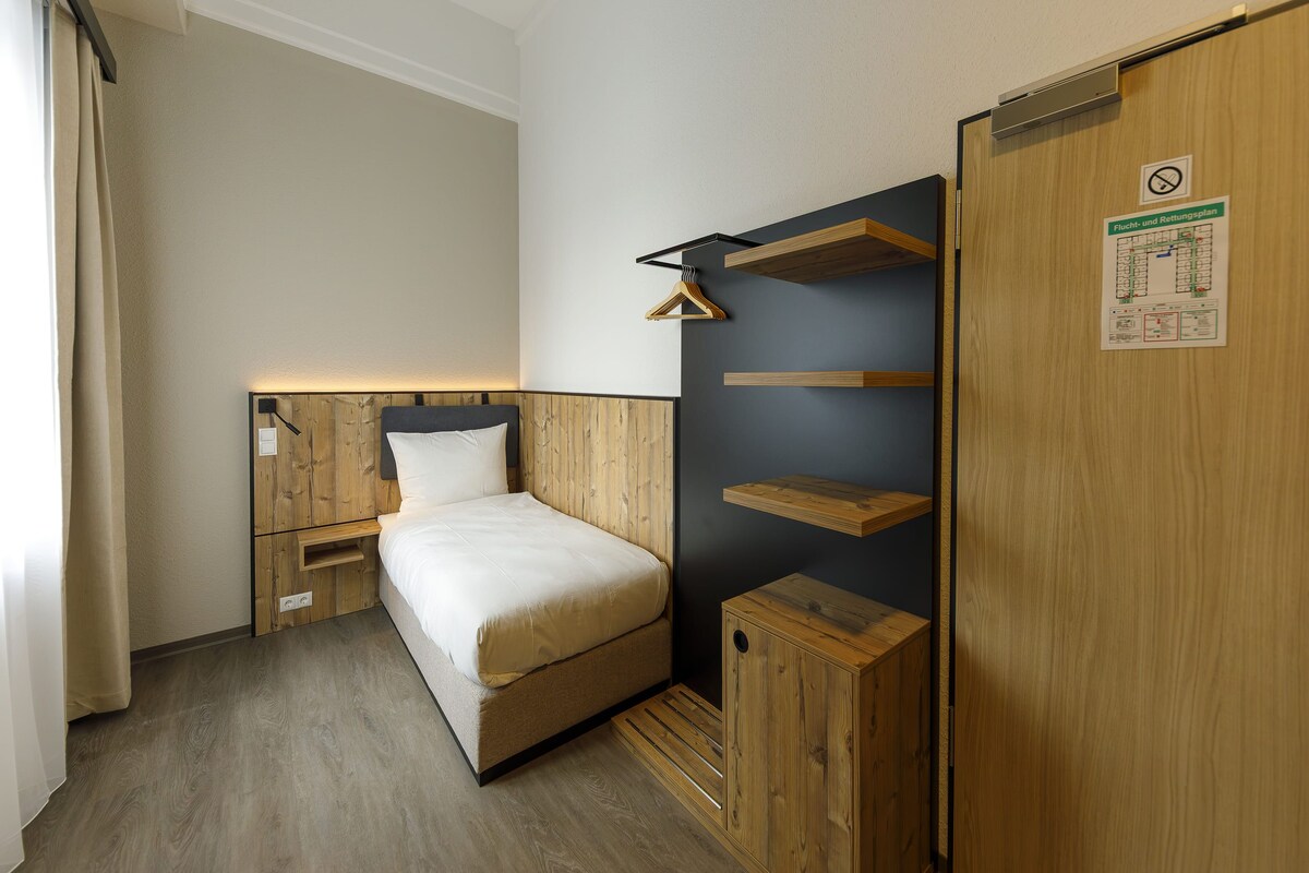 A hotel room with a bed, wooden wardrobe, and a wall-mounted shelf unit.