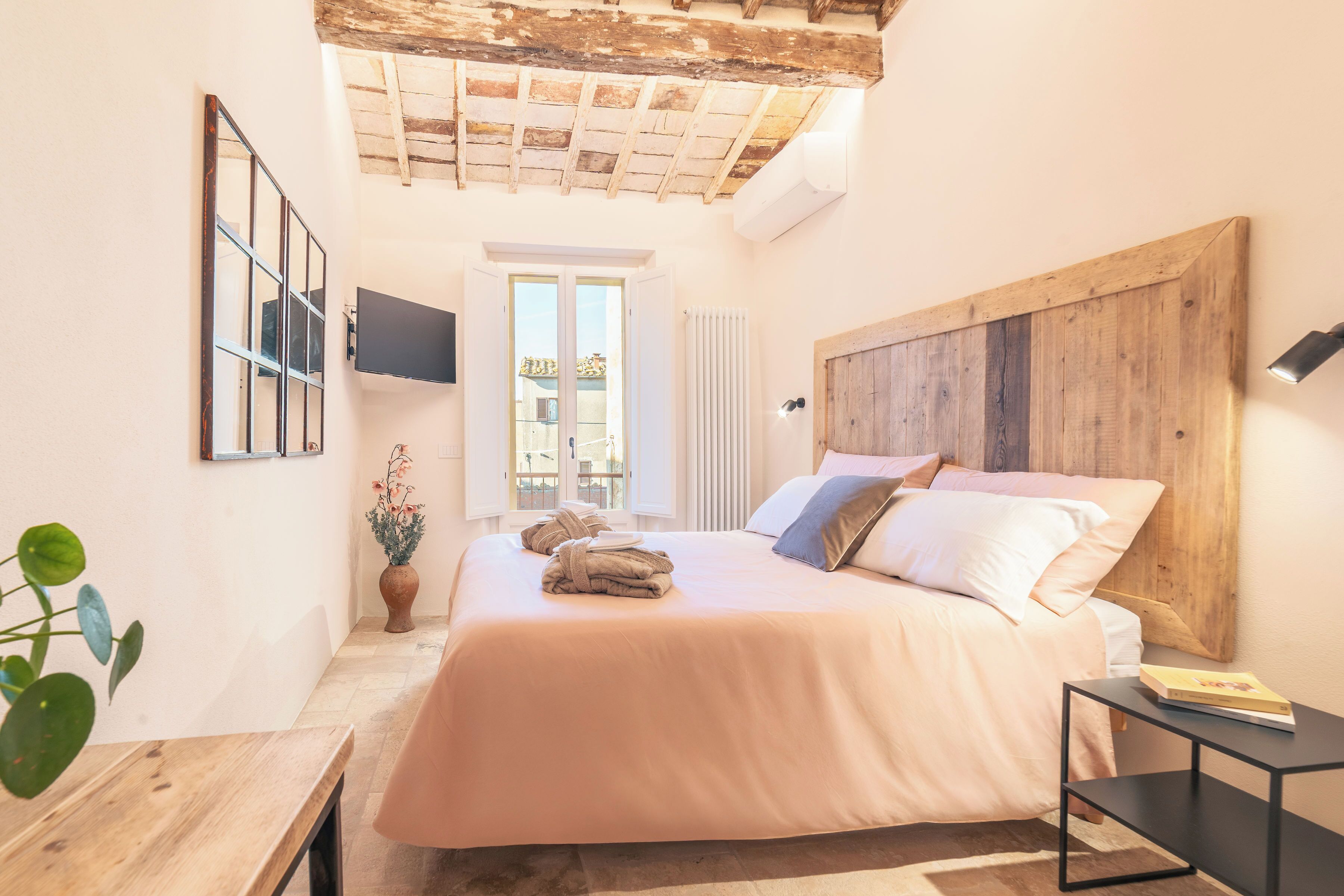 Ein Schlafzimmer mit Bett, Fernseher, Holz-Kopfstück, Pflanze und Fenster.