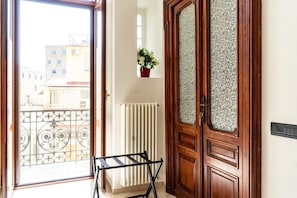 Um quarto com porta de madeira, radiador, janela com varanda e uma planta pendurada.