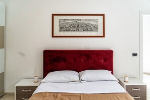 Een slaapkamer met een rood bekleed hoofdeinde, witte beddengoed en een zwart-wit stadsgezicht in een lijst aan de muur.