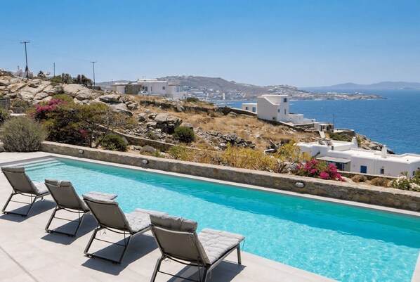A pool with lounge chairs, a view of the sea, and white buildings on a hillside. - Phos Chora - Six Bedroom Villa, Sleeps 12 (Mykonos)