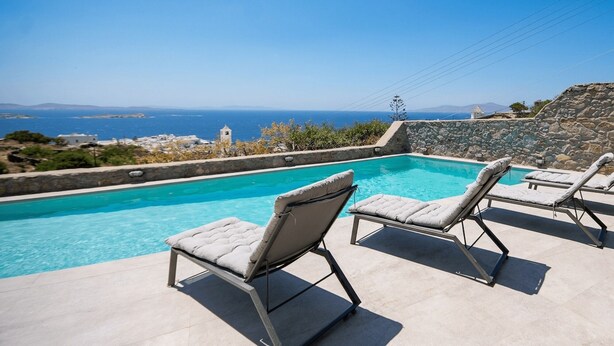 En pool med två solstolar, en stenmur och utsikt över havet och avlägsna byggnader.