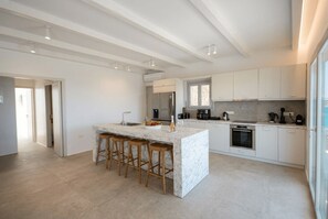 A modern kitchen with a marble island, wooden stools, stainless steel appliances, and white cabinetry. - Phos Chora - Six Bedroom Villa, Sleeps 12 (Mykonos)