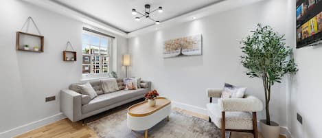 Una sala moderna con un sofá gris, un sillón blanco, una mesa de centro y un ventanal grande.