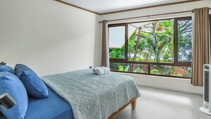 A bedroom with a bed, a window with a view of greenery, and a standing fan. - Relaxing Beachfront vacation in Costa Rica with a pool, overlooking the ocean! (Puntarenas Province)