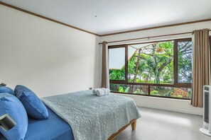 Ein Schlafzimmer mit einem Bett, einem Fenster mit Blick auf Grünflächen und einem Standventilator.