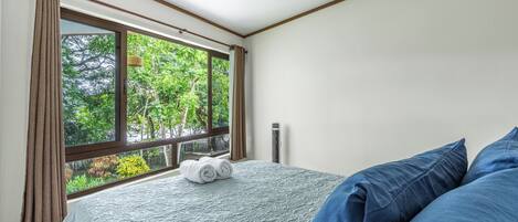 A bedroom with a large window, a bed with blue bedding, and a view of greenery outside.