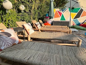 Eine Holzterrasse mit Liegestühlen und einem Tisch, umgeben von viel Grün und einer farbenfrohen Wand.