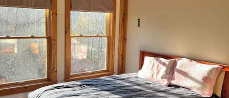 Un dormitorio con cama de madera, dos ventanas con persianas y una pared lisa.