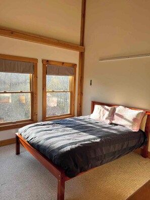 A bedroom with a wooden bed, two windows with blinds, and a plain wall.
