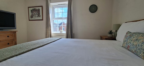 Une chambre à coucher avec un lit, un téléviseur posé sur une commode, une horloge au mur et une fenêtre avec des rideaux.
