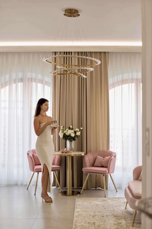 Eine Frau in einem weißen Kleid steht in einem Raum mit einem modernen Kronleuchter, zwei rosafarbenen Stühlen, einem runden Tisch mit einer Blumenvase und transparenten Vorhängen.
