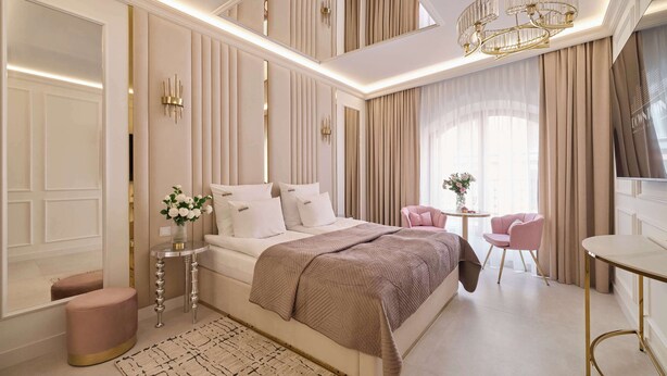 Een slaapkamer met een groot bed, een kroonluchter, een klein tafeltje met bloemen en een roze stoel.