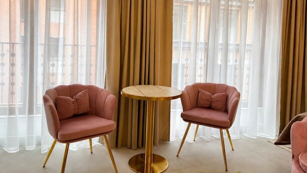 Ein Zimmer mit zwei rosafarbenen Sesseln und einem runden Tisch, großen Fenstern mit transparenten Vorhängen und Blick auf Gebäude.
