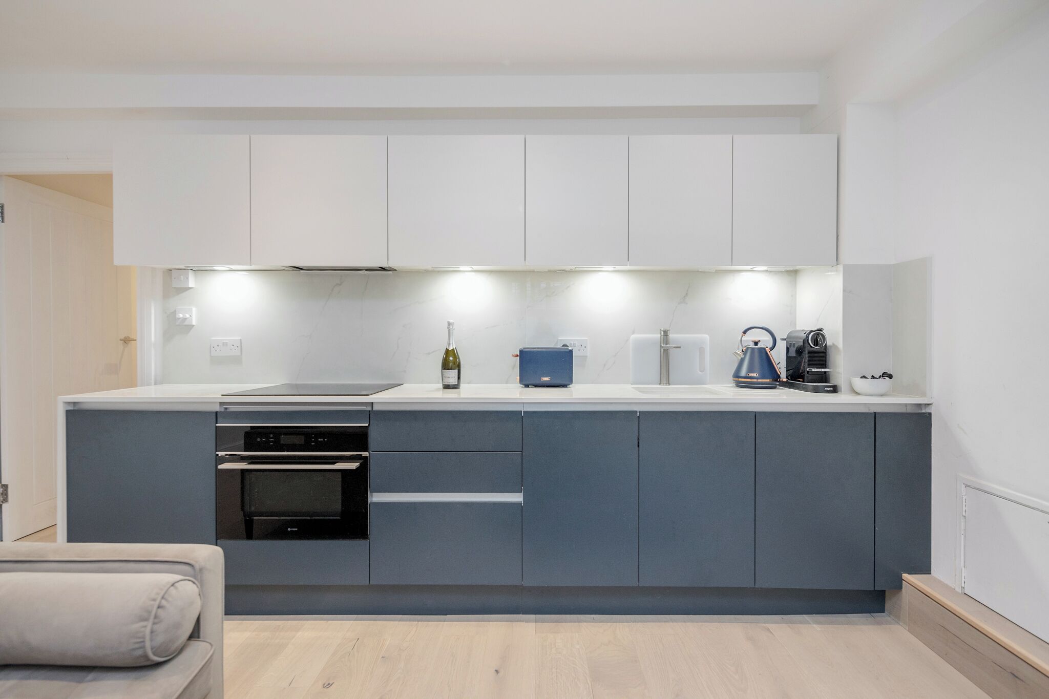 Eine moderne Küche mit grauen Schränken, einem eingebauten Backofen und einer Arbeitsfläche mit einer Weinflasche, einer Kaffeemaschine und einem Toaster.