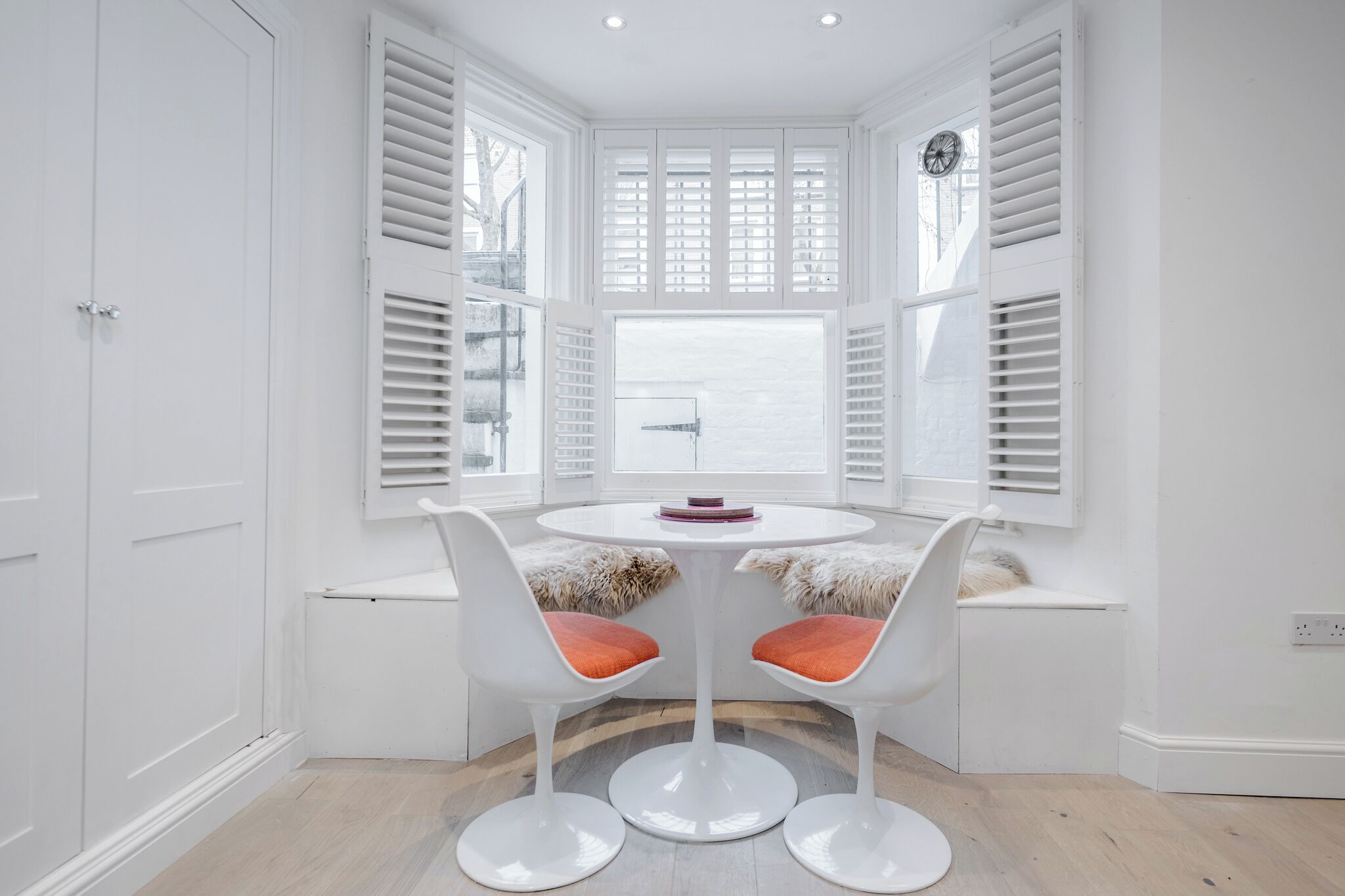 A corner room with a round table and two chairs, a window with shutters, and a white wardrobe.