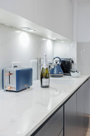 Uma cozinha moderna com bancada branca, torradeira azul, uma garrafa de Prosecco e chaleira.