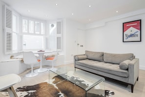 Uma sala moderna com uma mesa de centro de vidro, um sofá cinzento e um espaço de jantar com cadeiras brancas e uma mesa pequena.