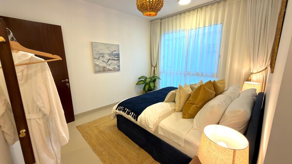 A bedroom with a bed, a bedside lamp, a window with curtains, a painting on the wall, and a door.