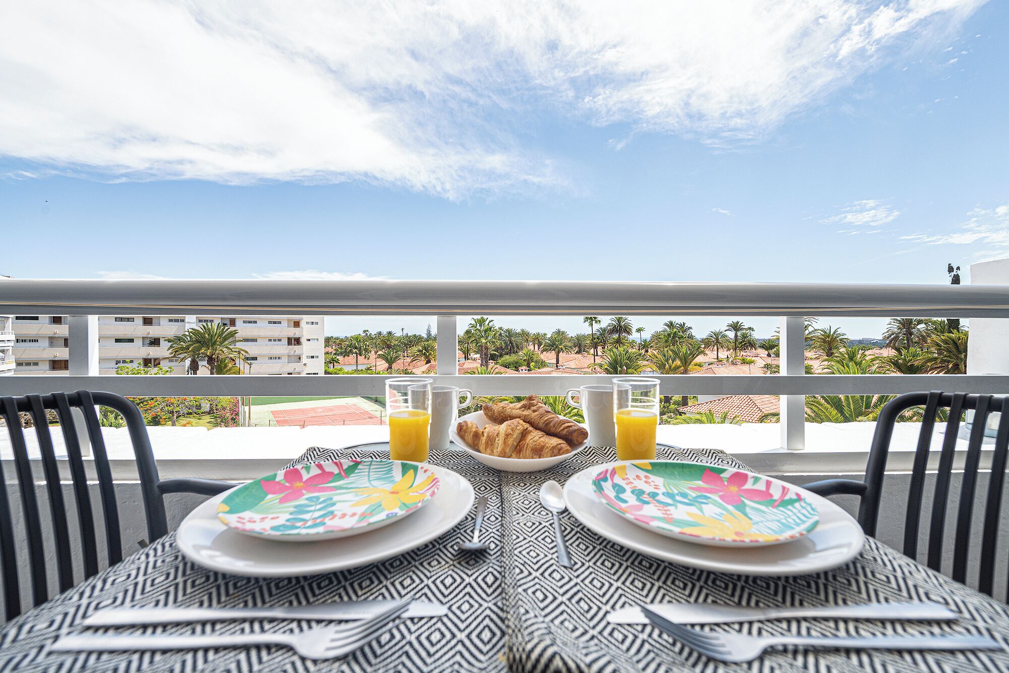 A table set for breakfast with croissants and orange juice, overlooking a tennis court and palm trees.