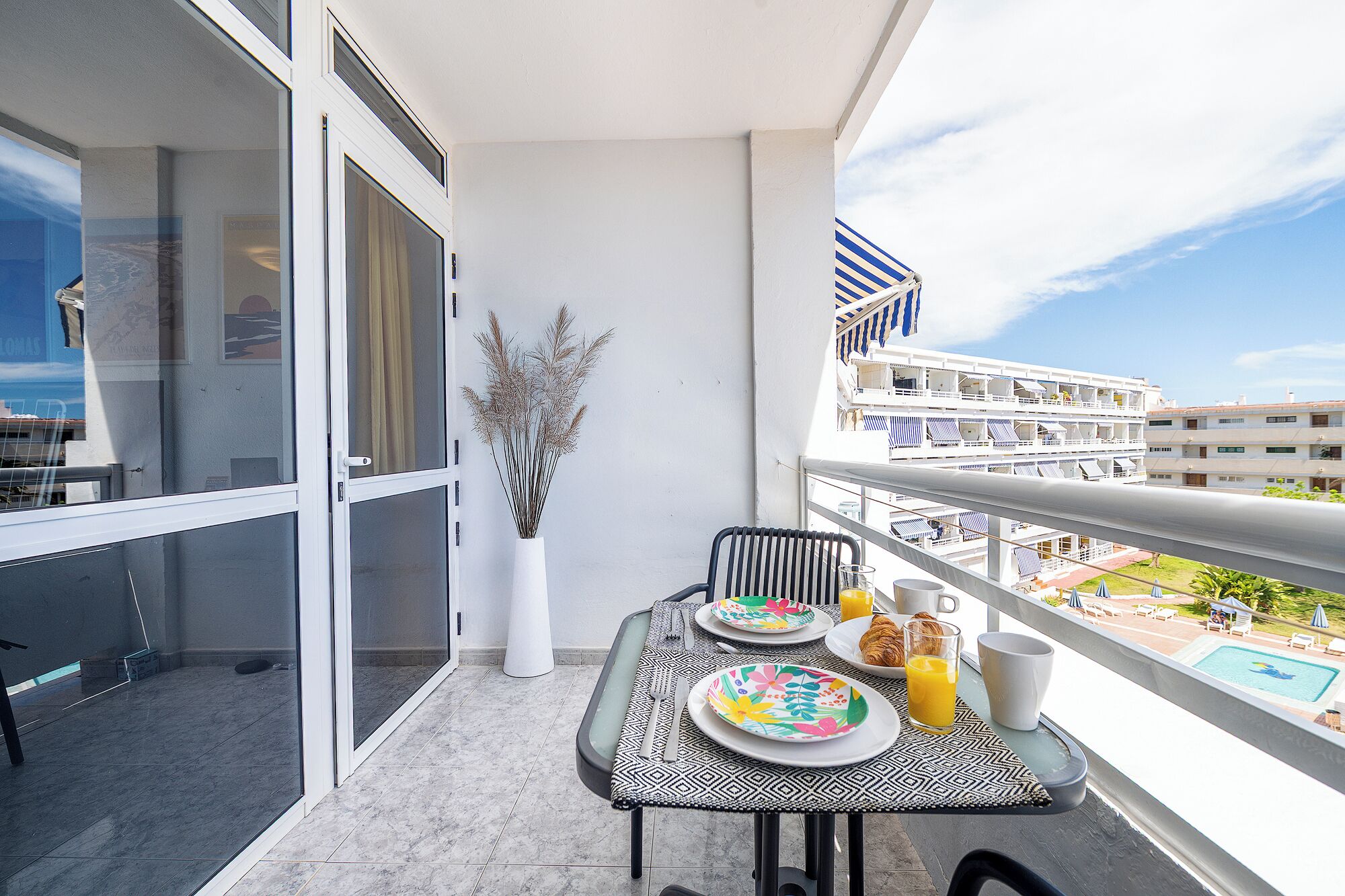 Een balkon met een ontbijttafel, een vaas met gedroogde planten en uitzicht op een cruiseschip en gebouwen.