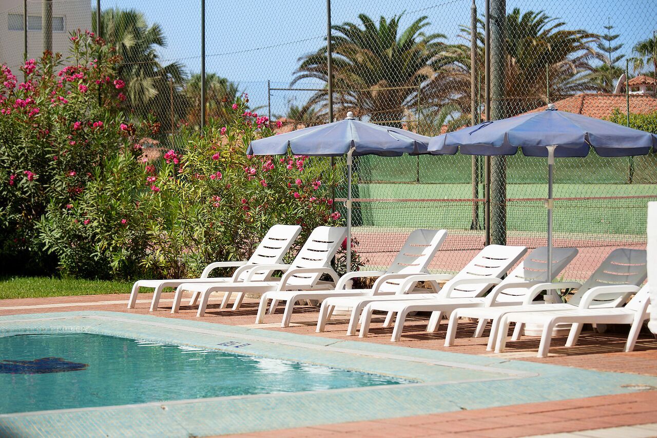 Una piscina con sillas de descanso blancas y sombrillas azules, rodeada de una valla y vegetación.