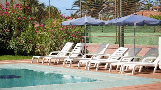 Una piscina con lettini bianchi e ombrelloni blu, circondata da una recinzione e vegetazione.