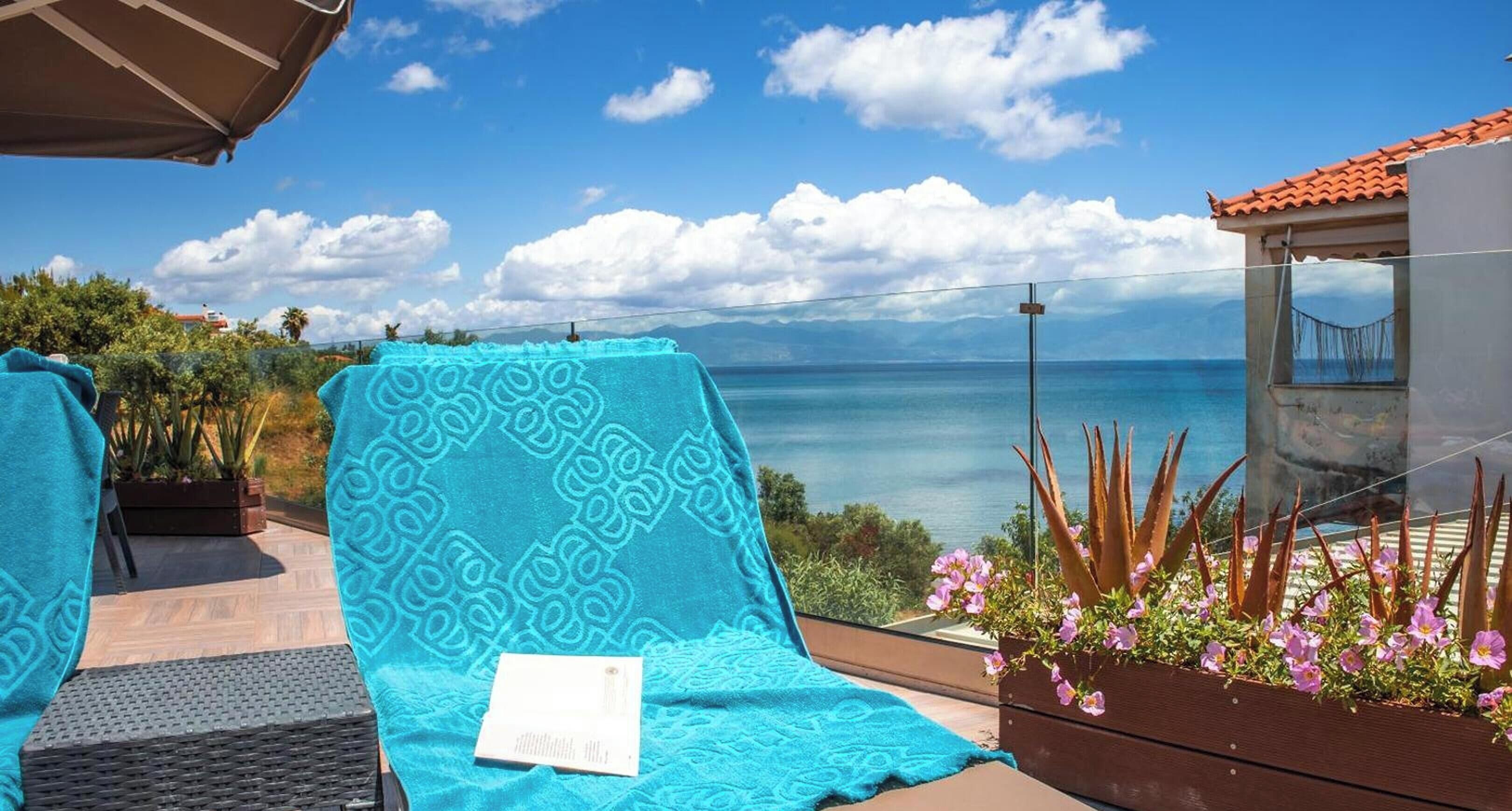 Un balcon avec vue sur la mer, une serviette bleue à motifs, une chaise et un jardinier en bois orné de fleurs.