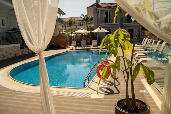 A swimming pool with a clear blue water, surrounded by lounge chairs and umbrellas, with a building in the background.