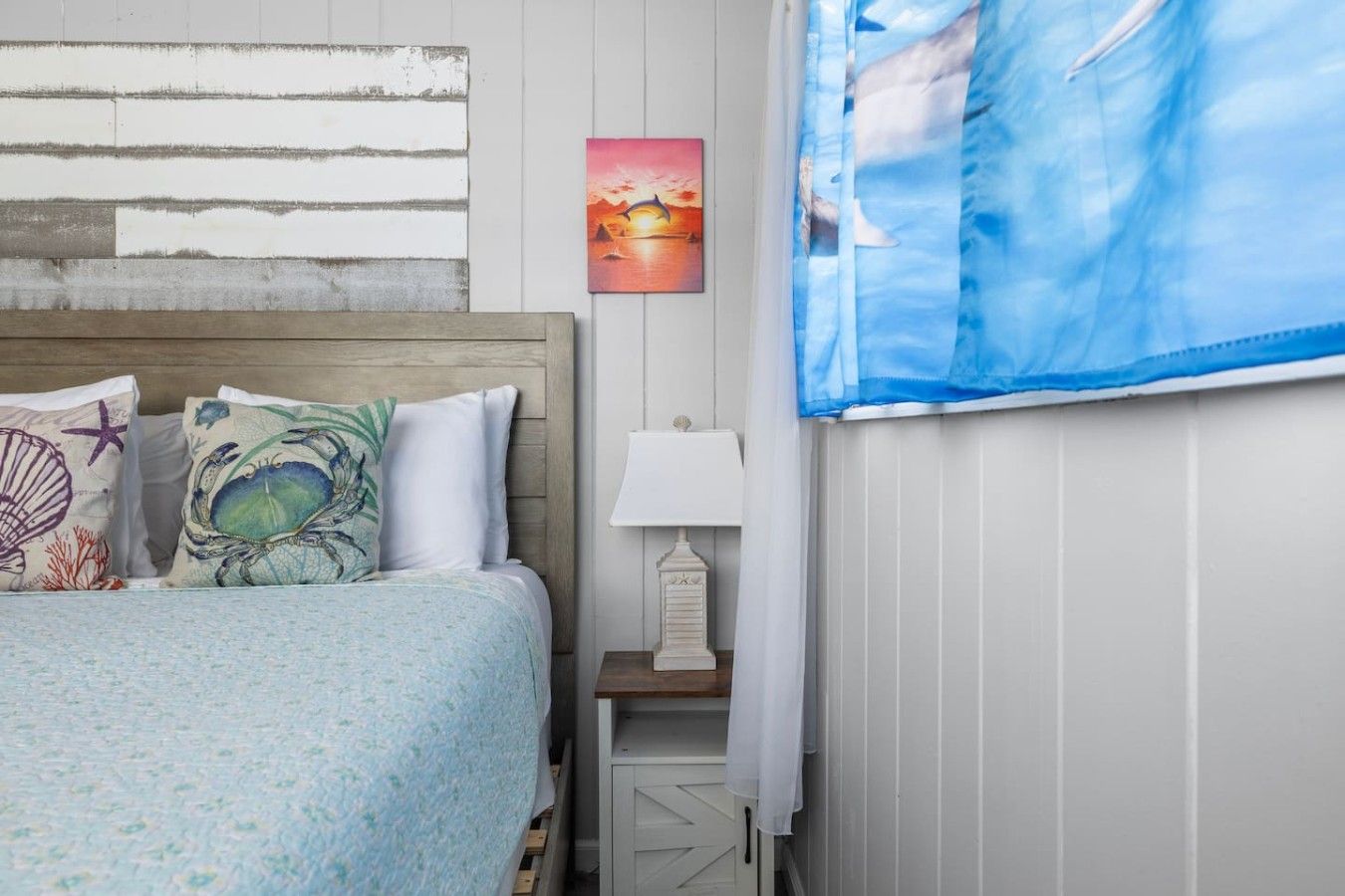 Een slaapkamer met een bed, een nachtkastje, een lamp, een schilderij en een raam met gordijnen.