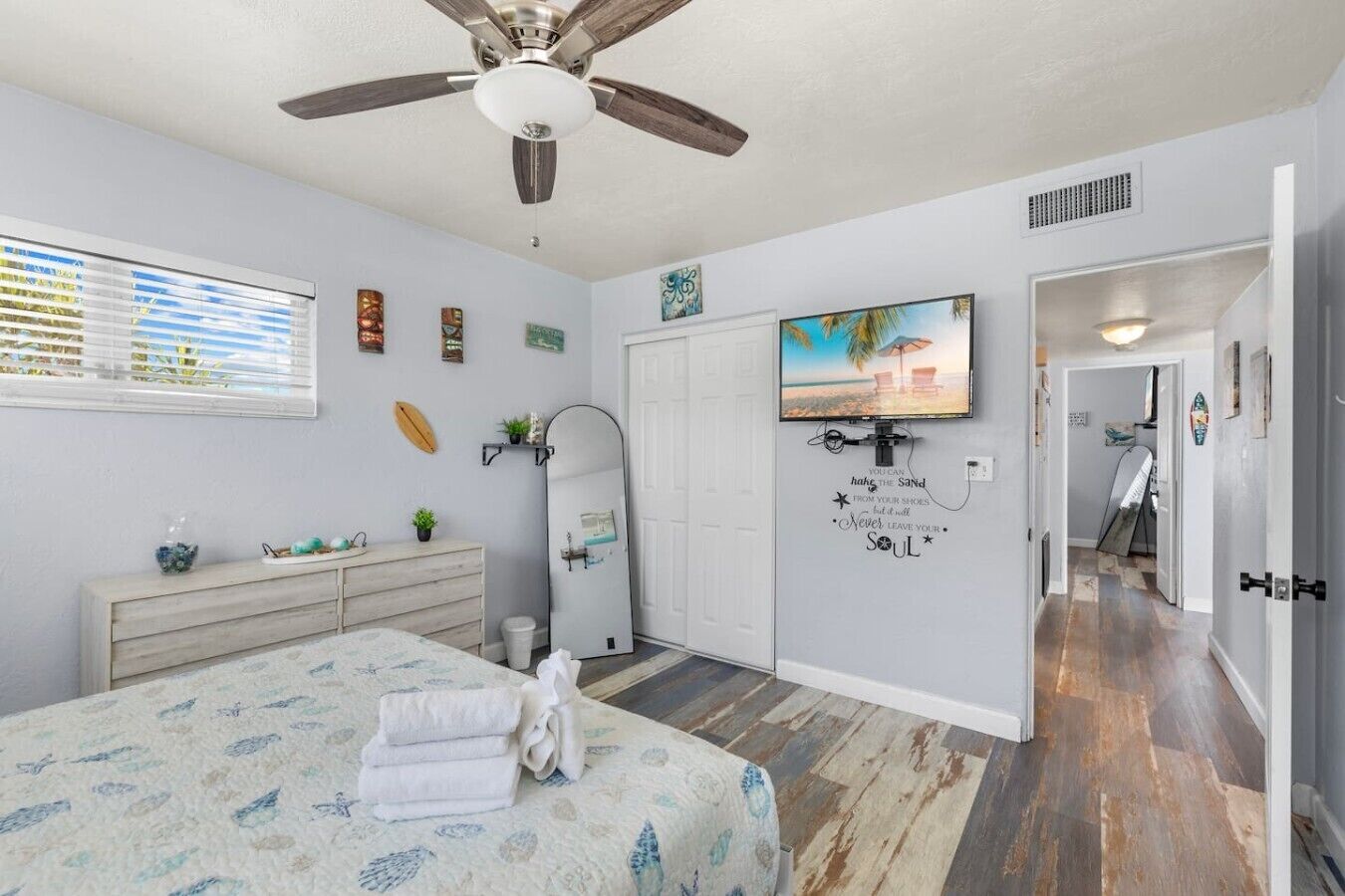 A bedroom with a bed, a ceiling fan, a large mirror, a TV, and a door leading to another room.