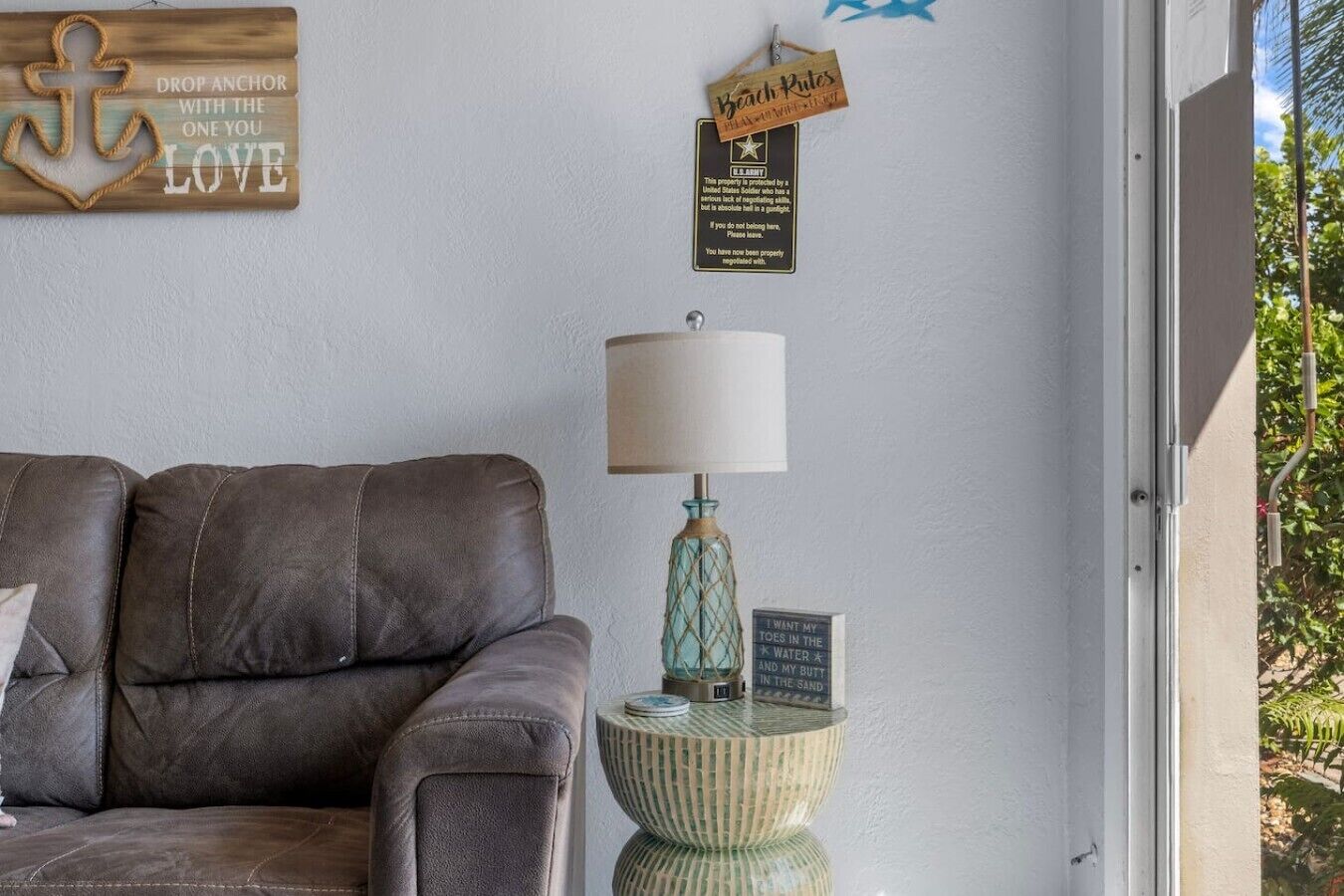 A room with a leather sofa, a lamp with a textured base, a small round table with a sign, and a wall with a wooden anchor decoration and a sign reading 'Beach Rules'.