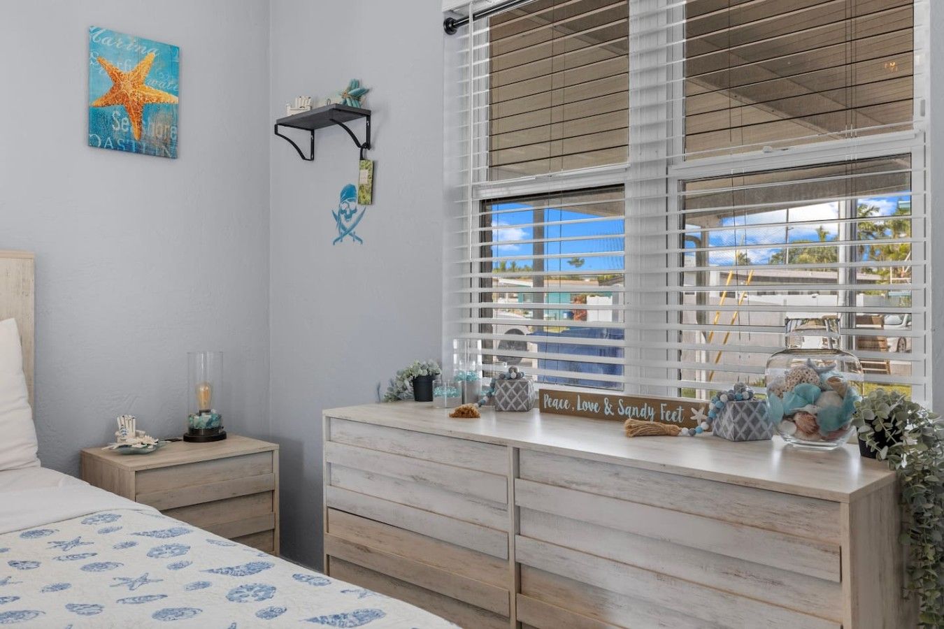 A bedroom with a bed, a dresser, a nightstand, a window with blinds, and a wall with a starfish artwork.