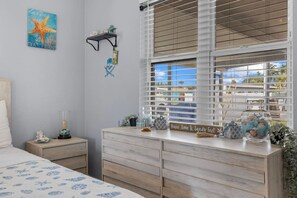 Une chambre à coucher comprenant un lit, une commode, une table de chevet, une fenêtre avec des stores et un mur orné d’une œuvre d’art représentant une étoile de mer.