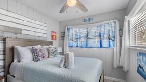 Een slaapkamer met een bed, een plafondventilator, een schilderij van vogels en een bordje met de tekst 'BEACH HOUSE'.
