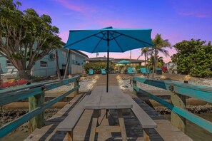Une terrasse en bois avec une table de pique-nique et un grand parasol, entourée de palmiers et d’un bâtiment doté d’une terrasse.