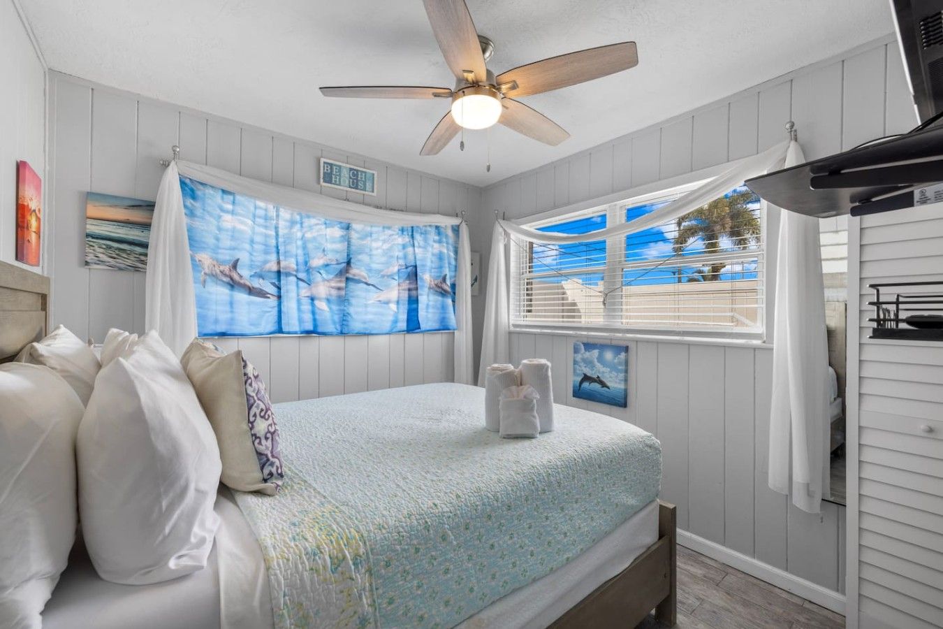 Quarto com cama, ventilador de teto, janela com vista e uma placa com a inscrição 'BEACH HOUSE'.