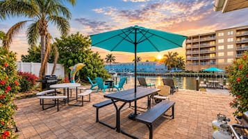 Une terrasse extérieure avec des tables et des chaises, un barbecue, et une vue sur une étendue d’eau et des bâtiments en arrière-plan.