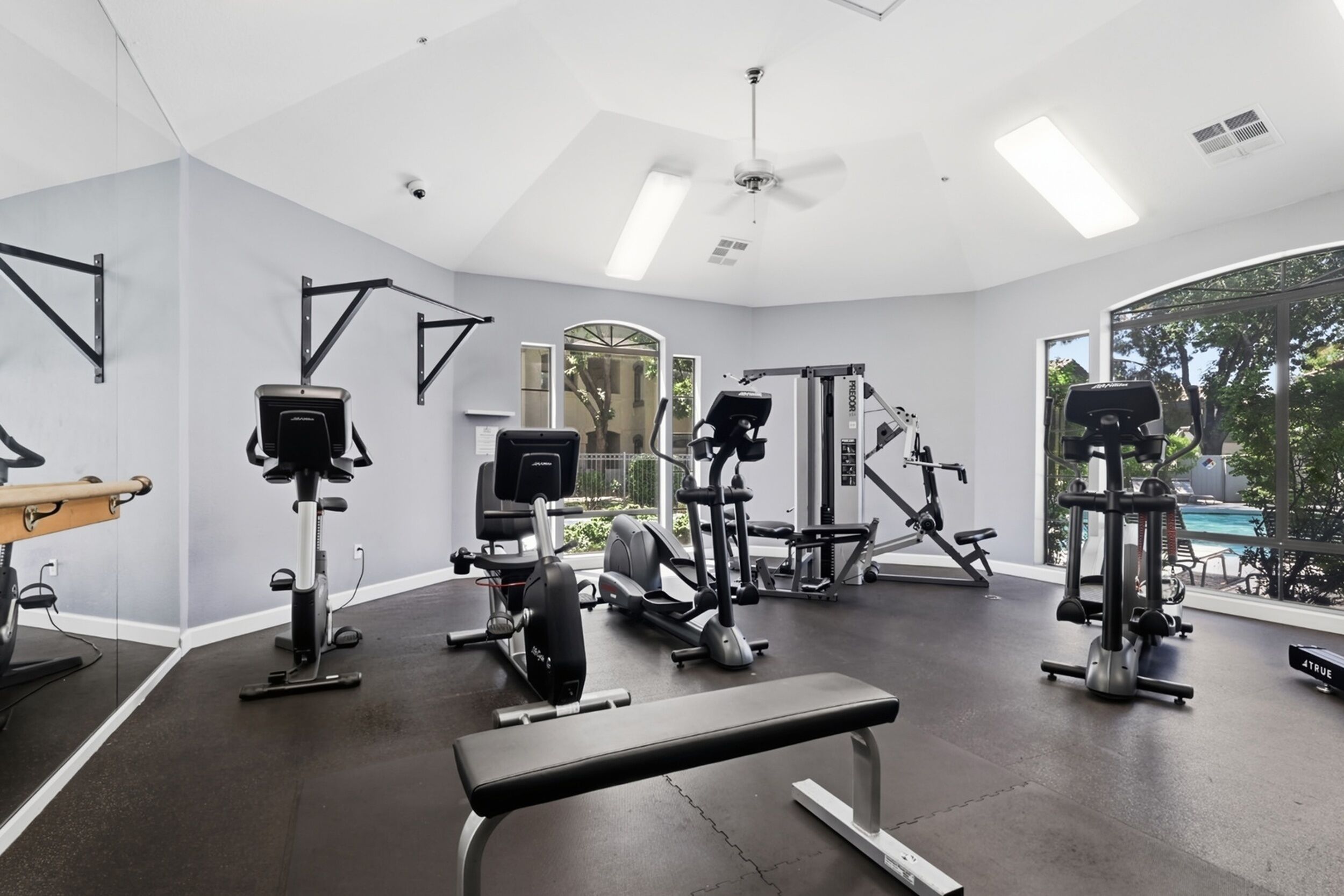 Une salle de sport équipée de divers appareils d’exercice, dotée d’un ventilateur de plafond et de grandes fenêtres.