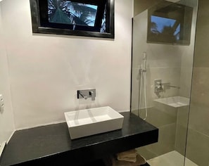 A bathroom with a black countertop, a white sink, a glass shower enclosure, and a mounted television.