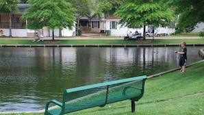 A green bench by a calm water body, a person fishing, and a row of houses in the background. - Ocean Lakes Beach House | Ocean View | Free Golf Cart | Sleeps 6 (Myrtle Beach)