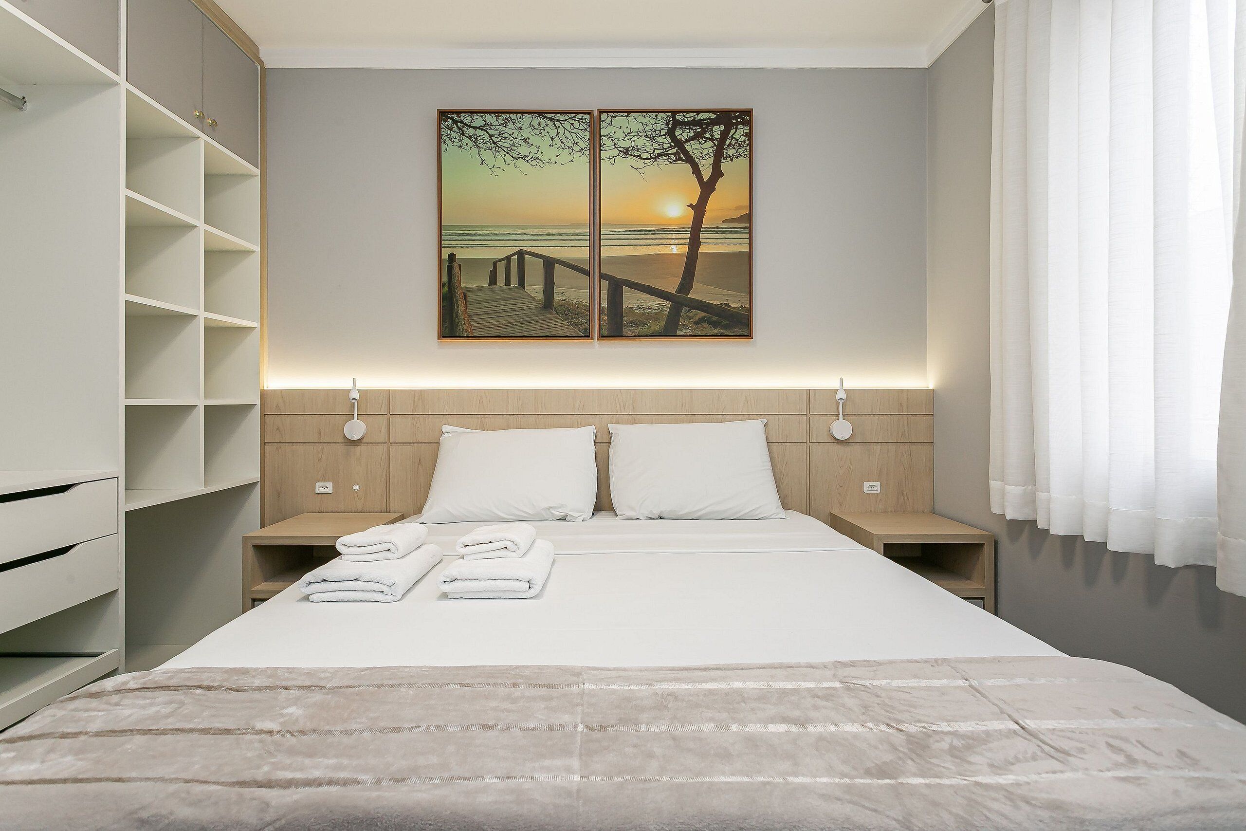 A modern bedroom with a bed, two nightstands, a wardrobe, and a painting of a sunset over a beach.